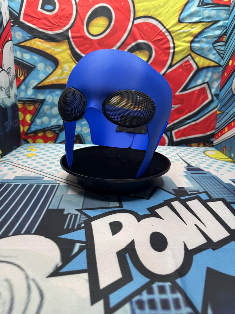 Puede incluir: Una m&aacute;scara de superh&eacute;roe azul con grandes aberturas para los ojos cubiertas de malla. La m&aacute;scara se exhibe sobre un soporte negro contra un fondo de gr&aacute;ficos de estilo c&oacute;mic. La m&aacute;scara tiene un acabado liso y est&aacute; dise&ntilde;ada para cubrir la parte superior de la cara.