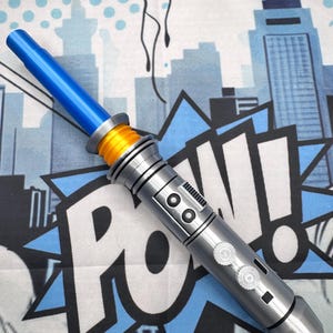May include: A blue and silver toy lightsaber with an orange accent. The lightsaber is set against a comic book-style background with the word "POW" and a cityscape. The lightsaber has a blue blade and a detailed silver handle.