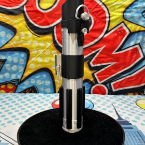 Może przedstawiać: Srebrna i czarna replika miecza świetlnego, z dopracowanym wzorem rękojeści i czarnymi akcentami. Miecza świetlnego jest zamontowany na czarnej, okrągłej podstawie. Tło to kolorowy wz&oacute;r w stylu komiksowym.