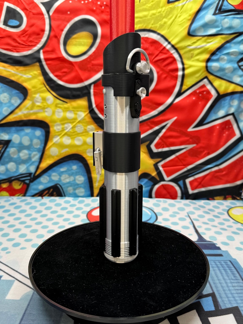 Może przedstawiać: Srebrna i czarna replika miecza świetlnego, z dopracowanym wzorem rękojeści i czarnymi akcentami. Miecza świetlnego jest zamontowany na czarnej, okrągłej podstawie. Tło to kolorowy wz&oacute;r w stylu komiksowym.