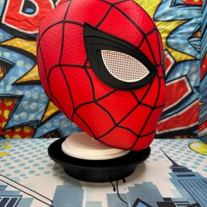 May include: A red Spider-Man mask with black webbing and eye details. The mask is displayed on a black and white stand. The background features a colorful comic book-style design.