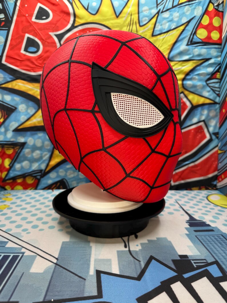 May include: A red Spider-Man mask with black webbing and eye details. The mask is displayed on a black and white stand. The background features a colorful comic book-style design.