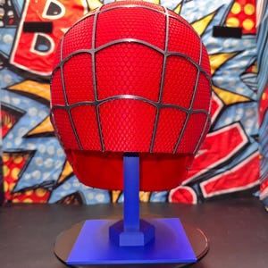 May include: A red Spider-Man helmet with a black web design, displayed on a blue stand. The helmet has a textured surface and is set against a colorful comic book-style backdrop. The stand is on a black surface.