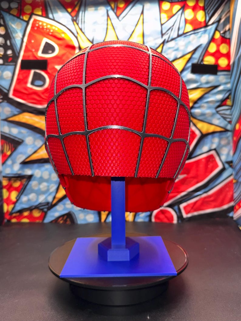 May include: A red Spider-Man helmet with a black web design, displayed on a blue stand. The helmet has a textured surface and is set against a colorful comic book-style backdrop. The stand is on a black surface.