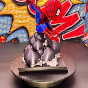 May include: A red, blue, and black Spider-Man figurine in a dynamic pose, perched atop a cluster of dark gray, spherical shapes. The figurine is on a round, black base. The background features a colorful comic book-style design.