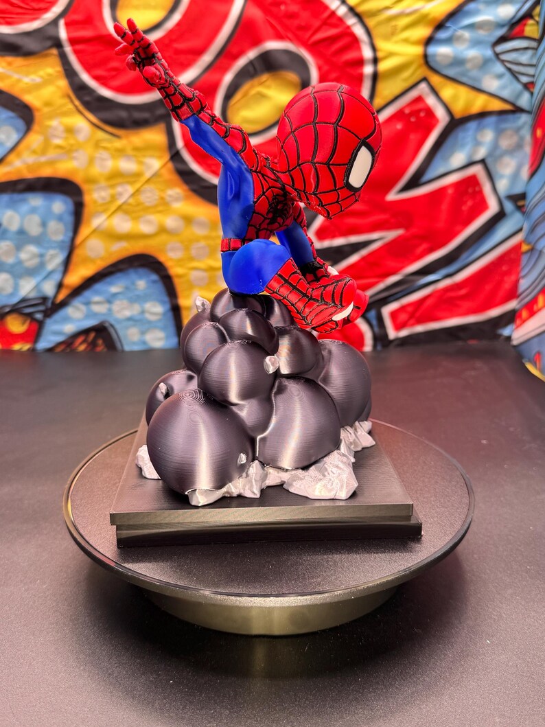 May include: A red, blue, and black Spider-Man figurine in a dynamic pose, perched atop a cluster of dark gray, spherical shapes. The figurine is on a round, black base. The background features a colorful comic book-style design.