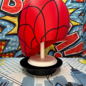 May include: A red Spider-Man mask with black webbing details, displayed on a white stand and a black base. The mask is a vibrant red with a textured surface. The background features a comic book-style design with bold colors and graphics.