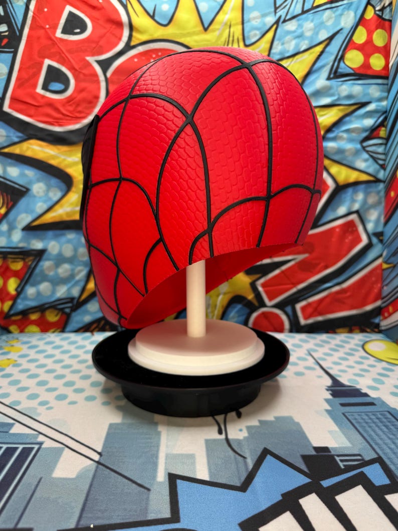 May include: A red Spider-Man mask with black webbing details, displayed on a white stand and a black base. The mask is a vibrant red with a textured surface. The background features a comic book-style design with bold colors and graphics.
