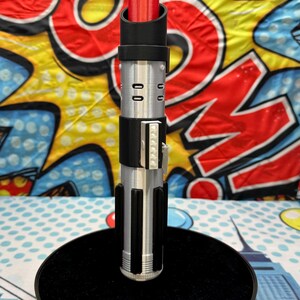 Może przedstawiać: Czerwony i srebrny rekwizyt miecza świetlnego z czarnymi akcentami, stojący na czarnej okrągłej podstawie. Miecz świetlny ma szczeg&oacute;łowy wz&oacute;r rękojeści ze srebrnymi i czarnymi elementami. Tło to kolorowy wz&oacute;r w stylu komiksowym.