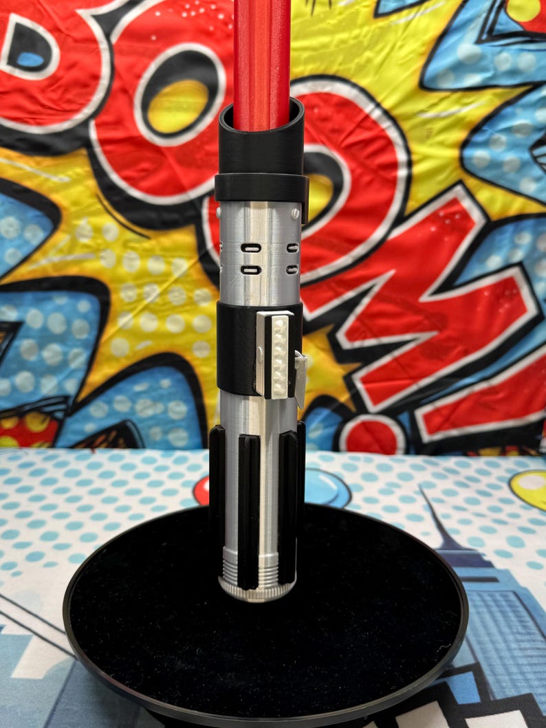 Może przedstawiać: Czerwony i srebrny rekwizyt miecza świetlnego z czarnymi akcentami, stojący na czarnej okrągłej podstawie. Miecz świetlny ma szczeg&oacute;łowy wz&oacute;r rękojeści ze srebrnymi i czarnymi elementami. Tło to kolorowy wz&oacute;r w stylu komiksowym.