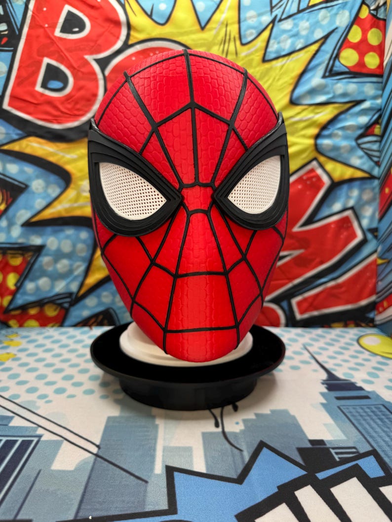 May include: A red and black Spider-Man mask with white eye mesh, displayed on a black stand. The mask features a textured surface and black web detailing. The background includes comic book-style graphics.