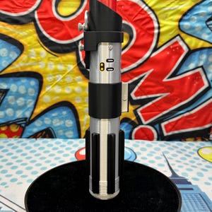 Może przedstawiać: Srebrna i czarna replika miecza świetlnego z czerwonym ostrzem, zamontowana na czarnej okrągłej podstawie. Rękojeść posiada szczeg&oacute;łowe elementy konstrukcyjne, w tym przyciski i czarny uchwyt. Tło to kolorowy wz&oacute;r w stylu komiksowym.