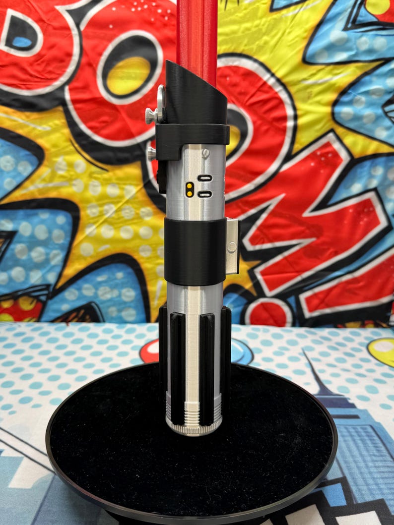 Może przedstawiać: Srebrna i czarna replika miecza świetlnego z czerwonym ostrzem, zamontowana na czarnej okrągłej podstawie. Rękojeść posiada szczeg&oacute;łowe elementy konstrukcyjne, w tym przyciski i czarny uchwyt. Tło to kolorowy wz&oacute;r w stylu komiksowym.