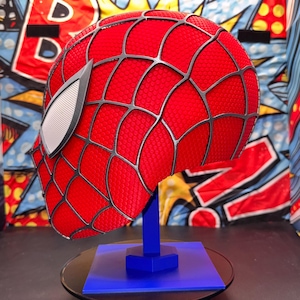 May include: A red Spider-Man mask with a black web pattern and white eye lenses, displayed on a blue stand. The mask is textured and has a detailed design, showcasing a comic book theme.