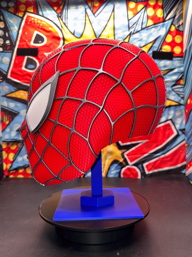 May include: A red Spider-Man mask with a black web pattern and white eye lenses, displayed on a blue stand. The mask is textured and has a detailed design, showcasing a comic book theme.