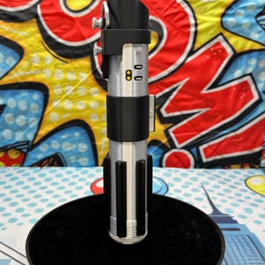 Może przedstawiać: Srebrno-czarna replika miecza świetlnego z czerwonym ostrzem, stojąca na czarnej, okrągłej podstawie. Rękojeść ma szczeg&oacute;łowe elementy, w tym przyciski i cylindryczną konstrukcję. Tło to kolorowy wz&oacute;r pop-art.