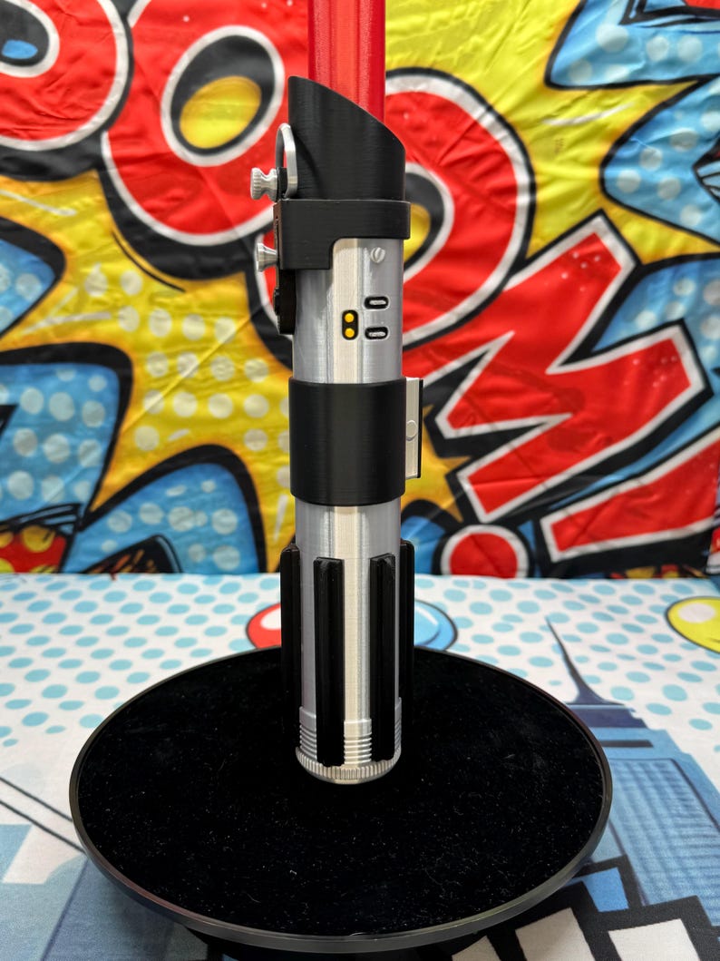 Może przedstawiać: Srebrno-czarna replika miecza świetlnego z czerwonym ostrzem, stojąca na czarnej, okrągłej podstawie. Rękojeść ma szczeg&oacute;łowe elementy, w tym przyciski i cylindryczną konstrukcję. Tło to kolorowy wz&oacute;r pop-art.