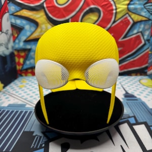 Puede incluir: Un casco amarillo texturizado con cubiertas oculares de malla blanca y finos soportes laterales amarillos. El casco se muestra sobre una base circular negra, con un fondo colorido de estilo c&oacute;mic.