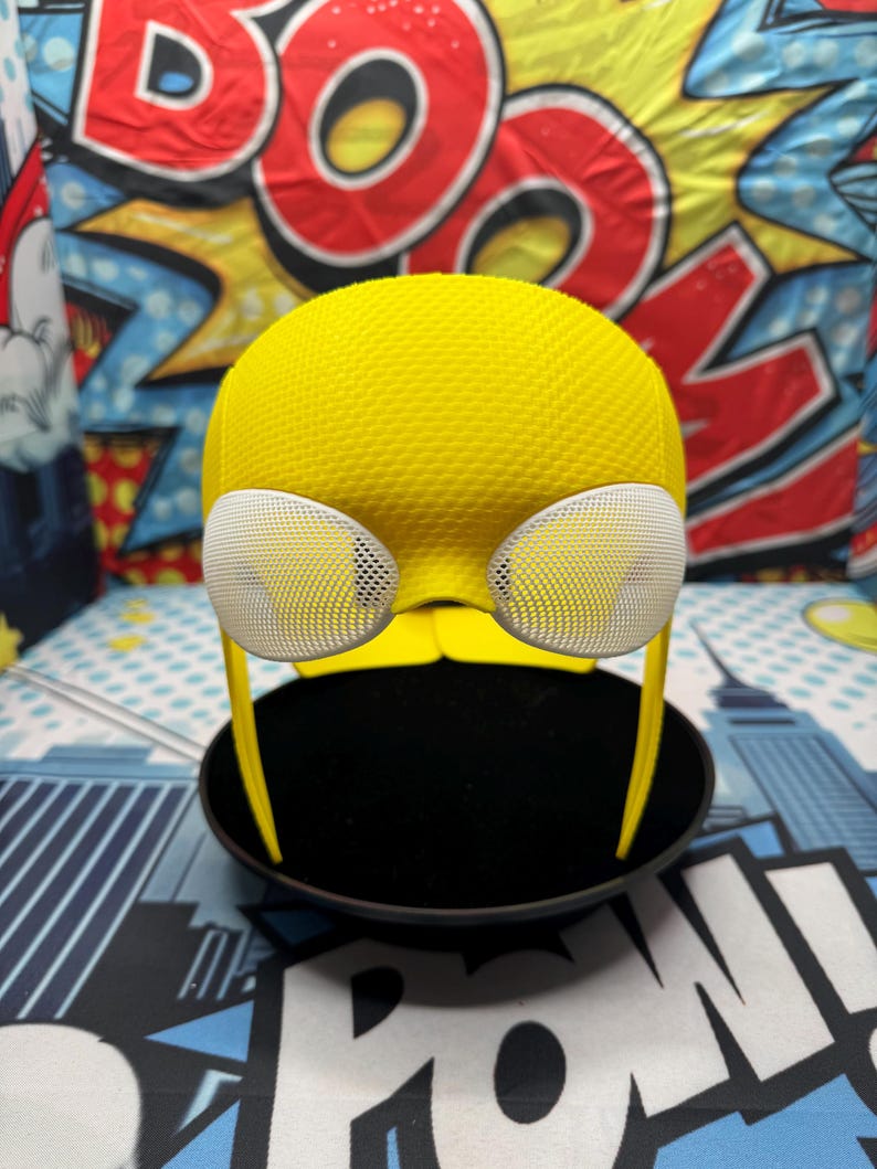 Puede incluir: Un casco amarillo texturizado con cubiertas oculares de malla blanca y finos soportes laterales amarillos. El casco se muestra sobre una base circular negra, con un fondo colorido de estilo c&oacute;mic.