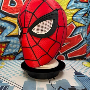 May include: A red and black Spider-Man mask with a web pattern and white eye lenses. The mask is displayed on a black stand against a colorful comic book-style background.
