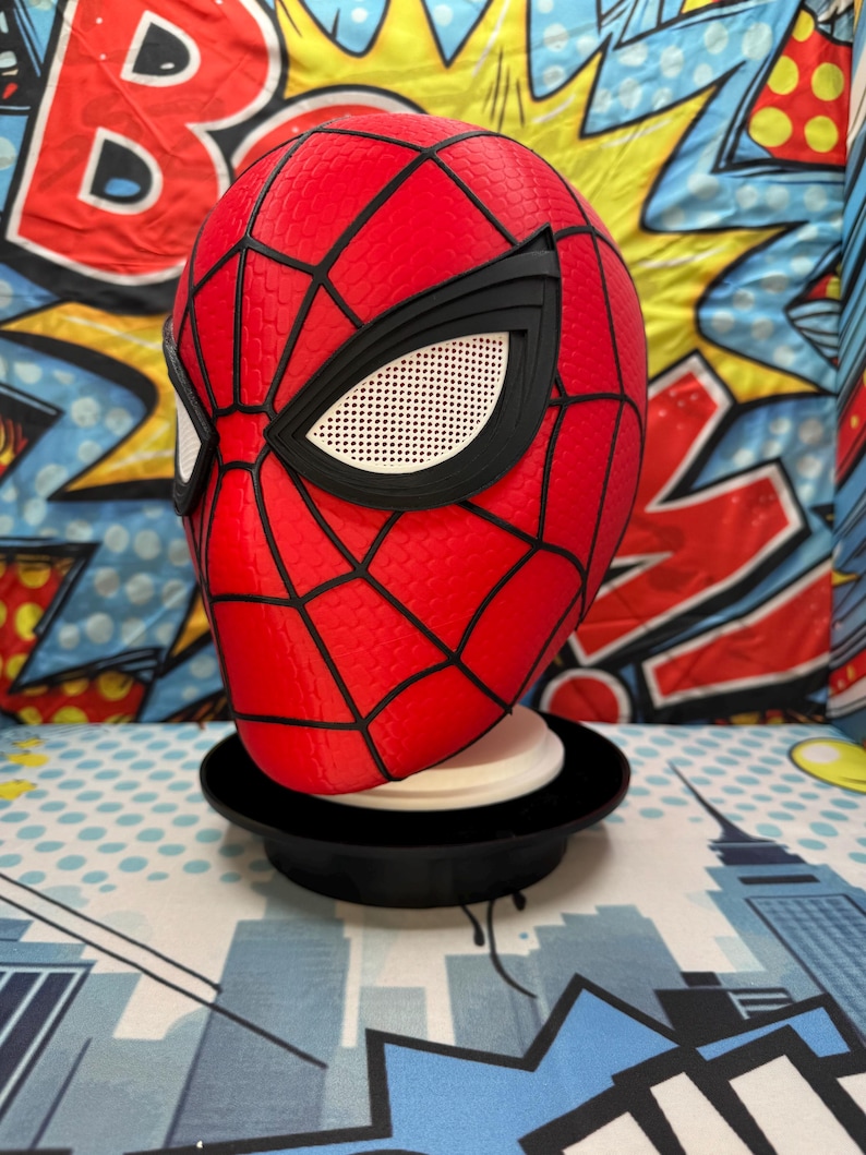 May include: A red and black Spider-Man mask with a web pattern and white eye lenses. The mask is displayed on a black stand against a colorful comic book-style background.