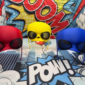 Puede incluir: Tres cascos coloridos de aspecto futurista en rojo, amarillo y azul. Cada casco tiene grandes cubiertas oculares de forma ovalada y un dise&ntilde;o suave y redondeado. El fondo presenta gr&aacute;ficos de estilo c&oacute;mic con la palabra "POW!"