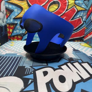 Puede incluir: Una m&aacute;scara azul con cubiertas oculares de malla negra se asienta sobre una base negra. La m&aacute;scara tiene un dise&ntilde;o curvo y est&aacute; hecha de un material liso. El fondo presenta gr&aacute;ficos de estilo c&oacute;mic con la palabra "POW".