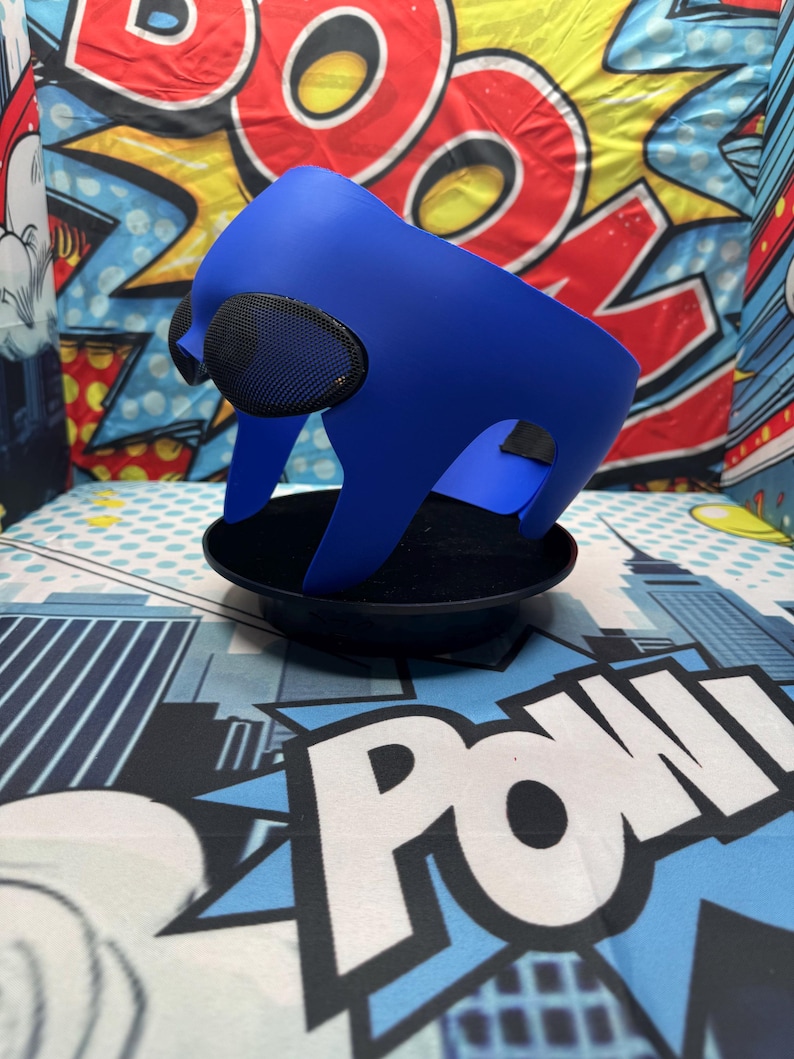 Puede incluir: Una m&aacute;scara azul con cubiertas oculares de malla negra se asienta sobre una base negra. La m&aacute;scara tiene un dise&ntilde;o curvo y est&aacute; hecha de un material liso. El fondo presenta gr&aacute;ficos de estilo c&oacute;mic con la palabra "POW".