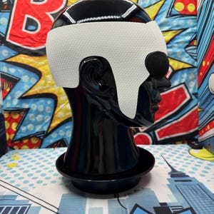 Puede incluir: Una cabeza de maniqu&iacute; negra con una cubierta para la cabeza blanca y negra. La cubierta para la cabeza tiene una banda blanca en la frente y alrededor de las orejas. El fondo es un dise&ntilde;o de arte pop colorido.