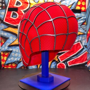 May include: A red Spider-Man helmet with a black web pattern, displayed on a blue stand. The helmet has a textured surface and is set against a colorful comic book-style background. The stand is square and sits on a black surface.