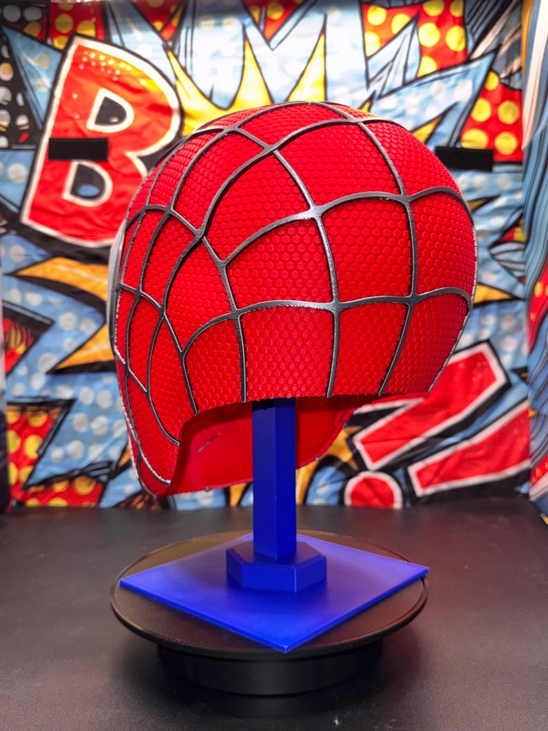 May include: A red Spider-Man helmet with a black web pattern, displayed on a blue stand. The helmet has a textured surface and is set against a colorful comic book-style background. The stand is square and sits on a black surface.