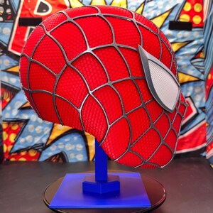May include: A red Spider-Man mask with black webbing and white eye lenses, displayed on a blue stand. The mask has a textured surface and is set against a colorful comic book-style backdrop.