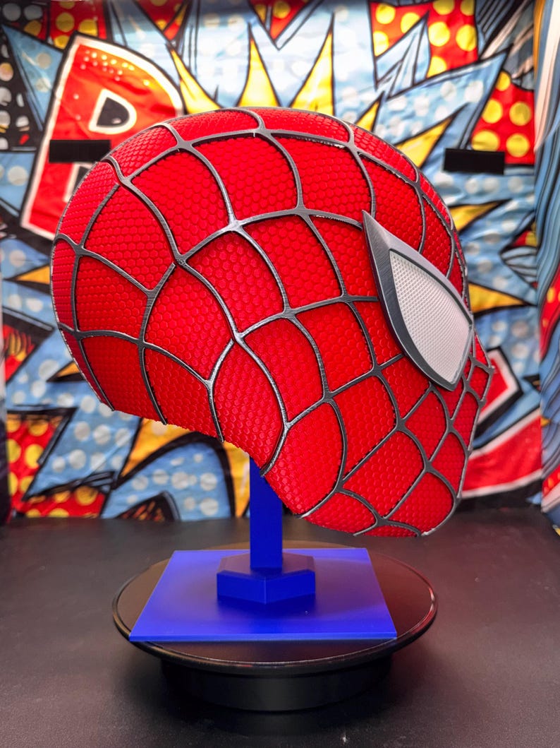 May include: A red Spider-Man mask with black webbing and white eye lenses, displayed on a blue stand. The mask has a textured surface and is set against a colorful comic book-style backdrop.