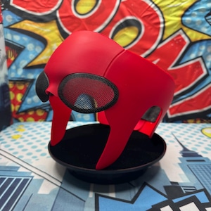 Puede incluir: Una m&aacute;scara roja brillante con cubiertas oculares de malla negra, colocada sobre un soporte de exhibici&oacute;n negro. La m&aacute;scara tiene un dise&ntilde;o angular &uacute;nico, probablemente un accesorio de disfraz o cosplay. El fondo presenta un dise&ntilde;o colorido estilo c&oacute;mic.