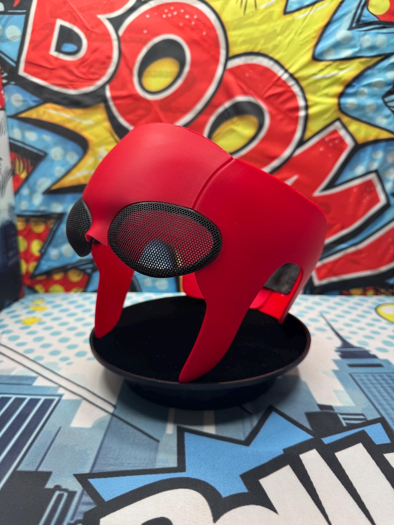 Puede incluir: Una m&aacute;scara roja brillante con cubiertas oculares de malla negra, colocada sobre un soporte de exhibici&oacute;n negro. La m&aacute;scara tiene un dise&ntilde;o angular &uacute;nico, probablemente un accesorio de disfraz o cosplay. El fondo presenta un dise&ntilde;o colorido estilo c&oacute;mic.