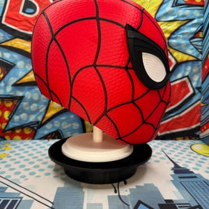 May include: A red Spider-Man mask with black webbing and white eye lenses. The mask is displayed on a white stand with a black base. The background features a colorful comic book-style design.