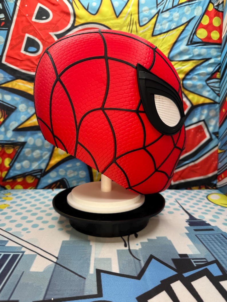 May include: A red Spider-Man mask with black webbing and white eye lenses. The mask is displayed on a white stand with a black base. The background features a colorful comic book-style design.
