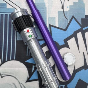 Puede incluir: Un sable de luz plateado y negro con botones rojos y verdes, un mango morado y una tapa plateada. El sable de luz est&aacute; sobre un fondo azul y blanco estilo c&oacute;mic.