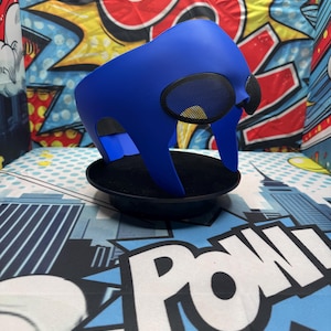 Puede incluir: Una llamativa m&aacute;scara azul con cubiertas oculares de malla, presentada sobre un fondo de gr&aacute;ficos estilo c&oacute;mic. La m&aacute;scara se exhibe sobre una base circular negra, resaltando su dise&ntilde;o y forma distintivos. La est&eacute;tica general sugiere un tema de superh&eacute;roe o cosplay.