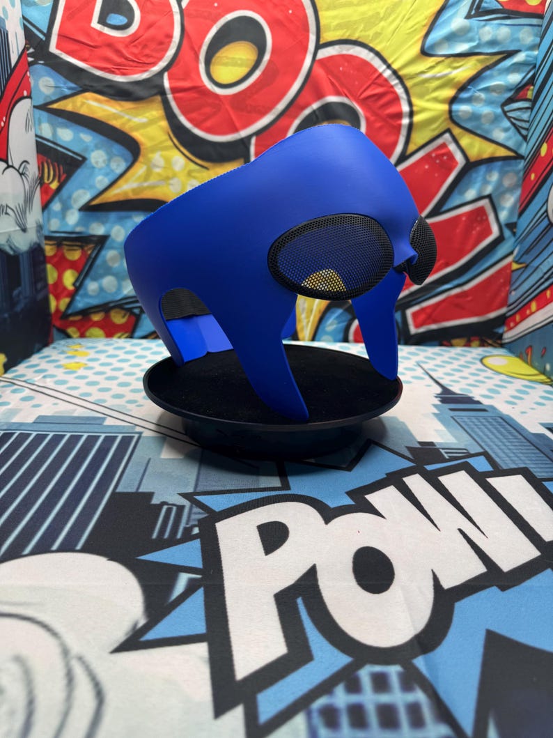 Puede incluir: Una llamativa m&aacute;scara azul con cubiertas oculares de malla, presentada sobre un fondo de gr&aacute;ficos estilo c&oacute;mic. La m&aacute;scara se exhibe sobre una base circular negra, resaltando su dise&ntilde;o y forma distintivos. La est&eacute;tica general sugiere un tema de superh&eacute;roe o cosplay.
