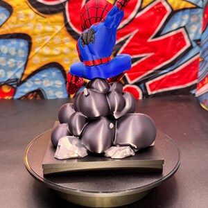 May include: A 3D-printed Spider-Man figurine in a dynamic pose, featuring a red and blue costume. The figure is perched atop a cluster of dark gray, rock-like structures, set on a black base. The background has a comic book style design.