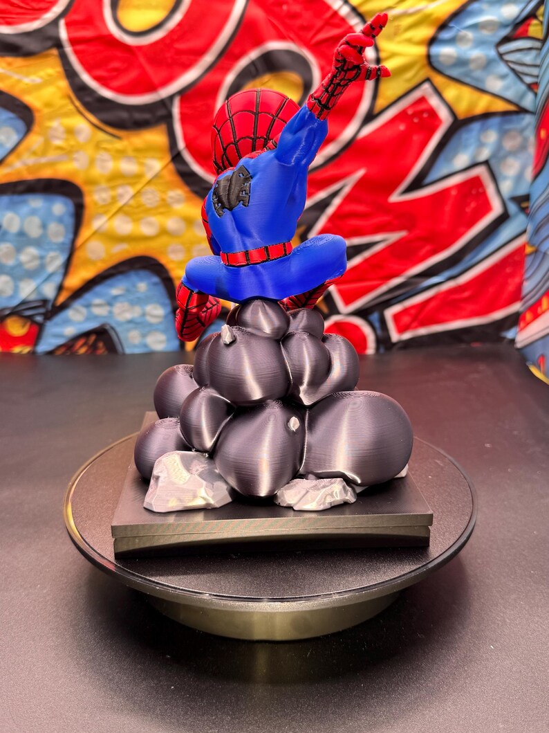 May include: A 3D-printed Spider-Man figurine in a dynamic pose, featuring a red and blue costume. The figure is perched atop a cluster of dark gray, rock-like structures, set on a black base. The background has a comic book style design.