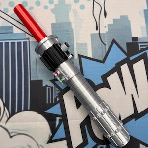 Puede incluir: Un sable de luz plateado y negro con una hoja roja. El mango presenta botones y un dise&ntilde;o acanalado. El sable de luz se encuentra sobre un fondo de estilo c&oacute;mic con siluetas de ciudades y bocadillos.