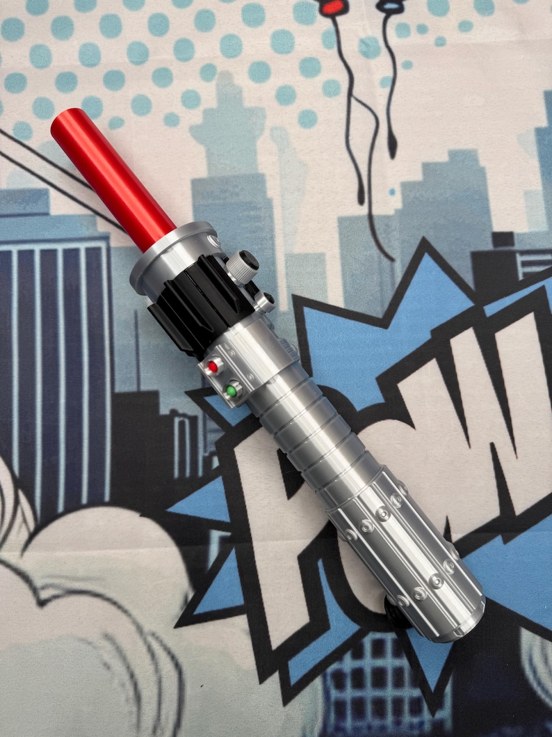 Puede incluir: Un sable de luz plateado y negro con una hoja roja. El mango presenta botones y un dise&ntilde;o acanalado. El sable de luz se encuentra sobre un fondo de estilo c&oacute;mic con siluetas de ciudades y bocadillos.