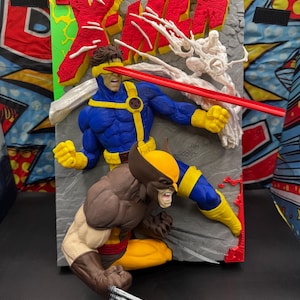 Op de afbeelding: Een 3D-muurkunstwerk met de Marvel Comics X-Men personages Cyclops en Wolverine. Cyclops draagt een blauw en geel pak met een rode optische straal, terwijl Wolverine een bruin en geel pak draagt met zilveren klauwen. De achtergrond is van grijze steen.