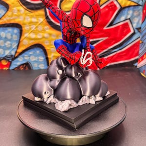 May include: A red, blue, and white Spider-Man figurine, perched atop a dark gray cloud-like structure. The figurine is on a black square base, set on a rotating platform. The background features a colorful comic book-style design.