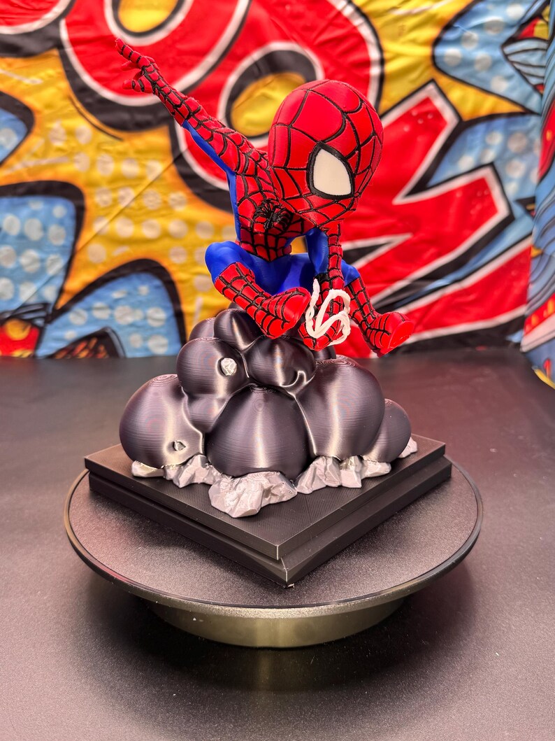 May include: A red, blue, and white Spider-Man figurine, perched atop a dark gray cloud-like structure. The figurine is on a black square base, set on a rotating platform. The background features a colorful comic book-style design.
