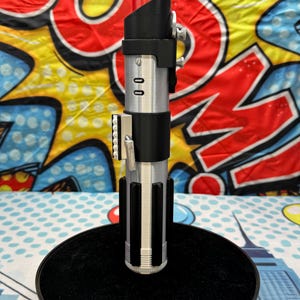 Może przedstawiać: Srebrno-czarna replika miecza świetlnego z czerwonym ostrzem, stojąca na czarnej, okrągłej podstawie. Rękojeść posiada szczeg&oacute;łowe elementy, w tym przyciski i uchwyt. Tło stanowi kolorowy wz&oacute;r pop-art.