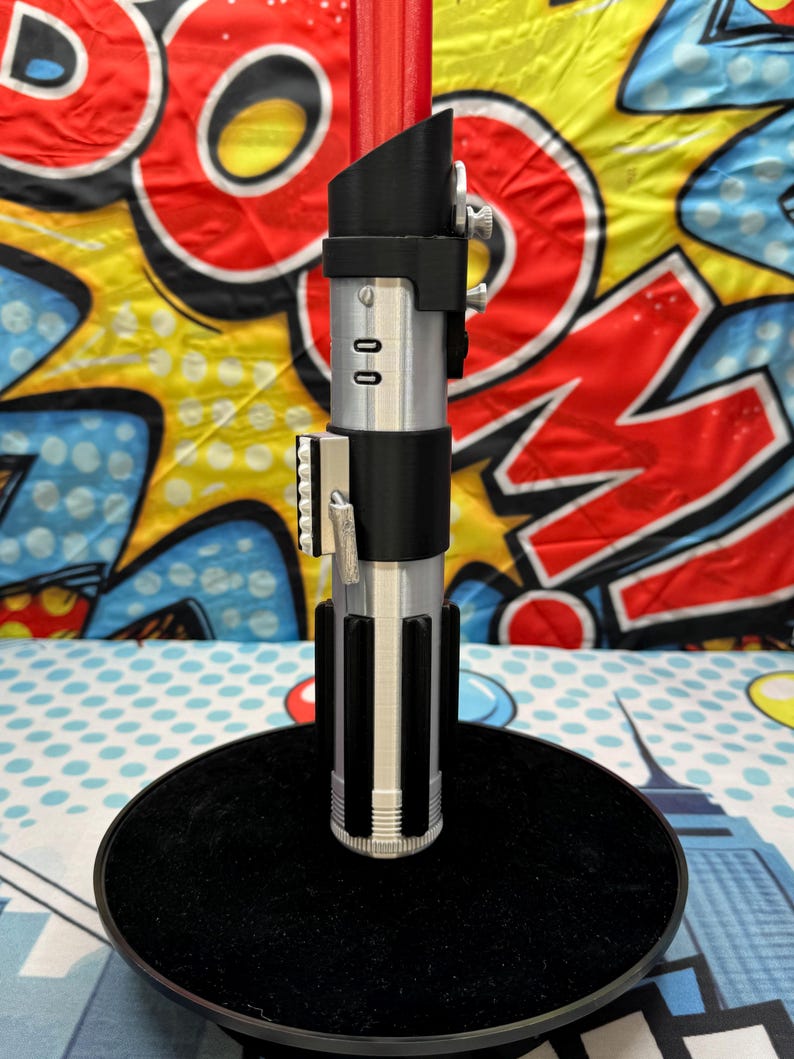 Może przedstawiać: Srebrno-czarna replika miecza świetlnego z czerwonym ostrzem, stojąca na czarnej, okrągłej podstawie. Rękojeść posiada szczeg&oacute;łowe elementy, w tym przyciski i uchwyt. Tło stanowi kolorowy wz&oacute;r pop-art.