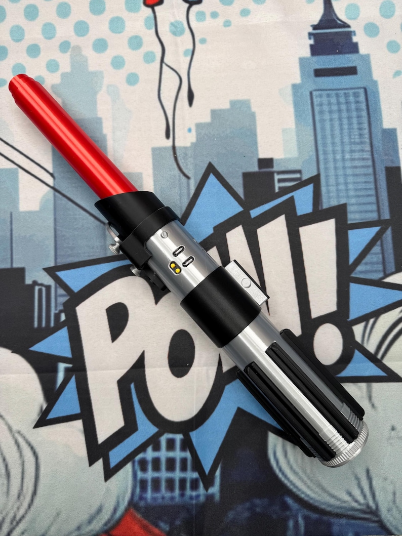 Może przedstawiać: Czerwony i srebrny rekwizyt miecza świetlnego z czarnym uchwytem. Miecz świetlny ma czerwone ostrze i srebrną rękojeść z czarnymi akcentami. Rekwizyt z serii Gwiezdne Wojny.